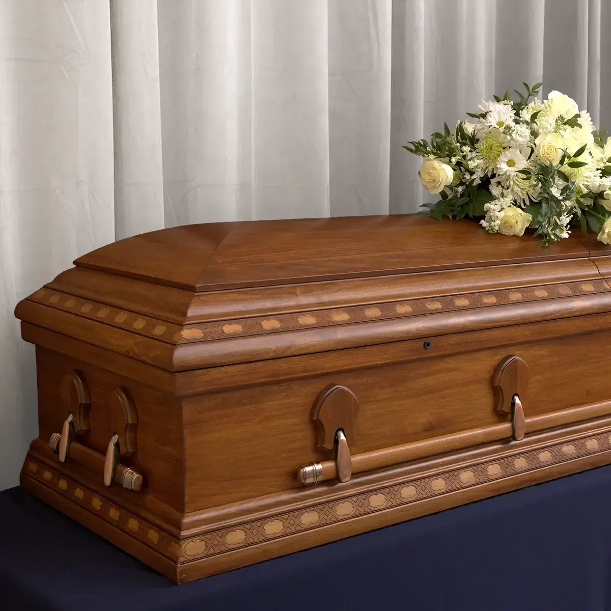 Wooden casket with brass handles, topped with white flowers, on a draped table