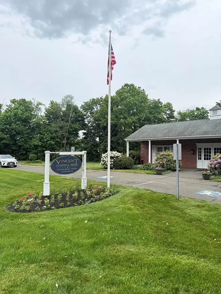 American flag beside a sign and brick building in a grassy landscaped area under cloudy skies