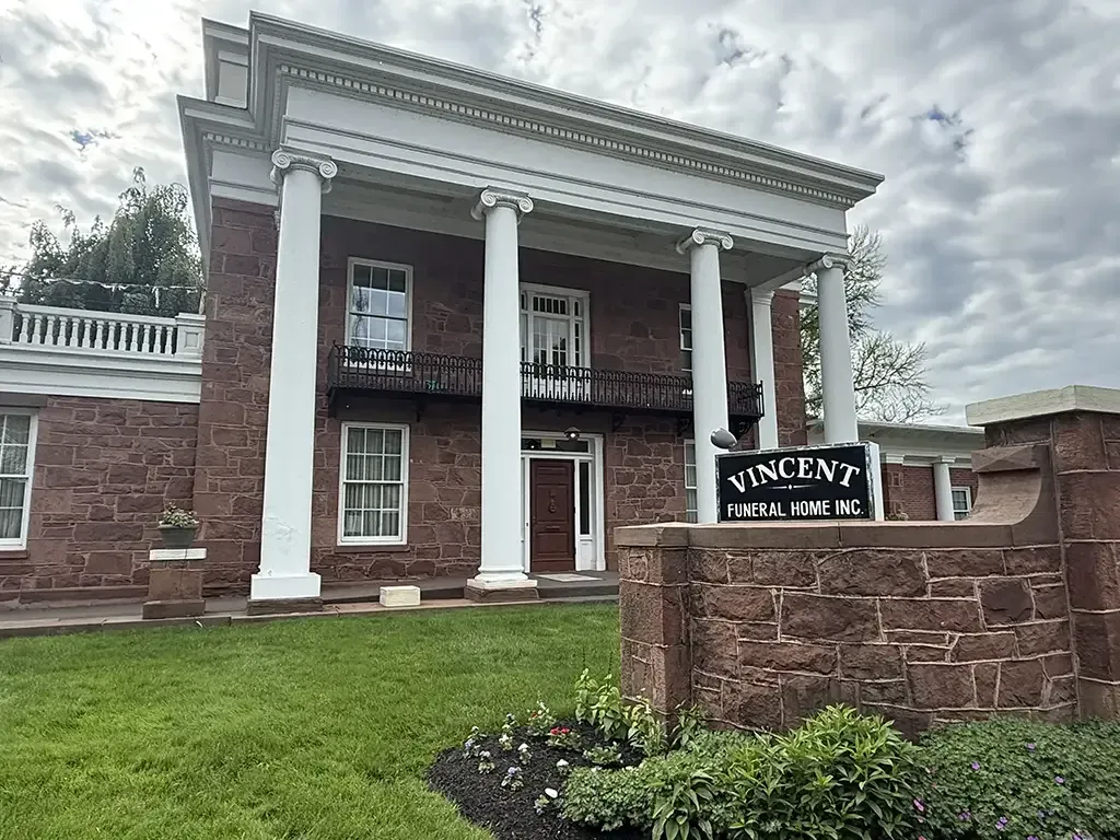 Historic brick building with white columns and a sign reading “Vincent Funeral Home” behind a stone wall.