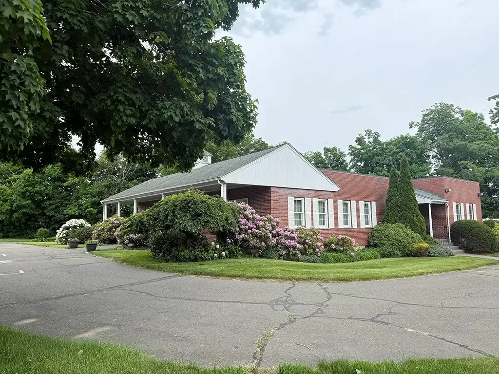 One-story brick building with white trim beside a parking lot and large trees
