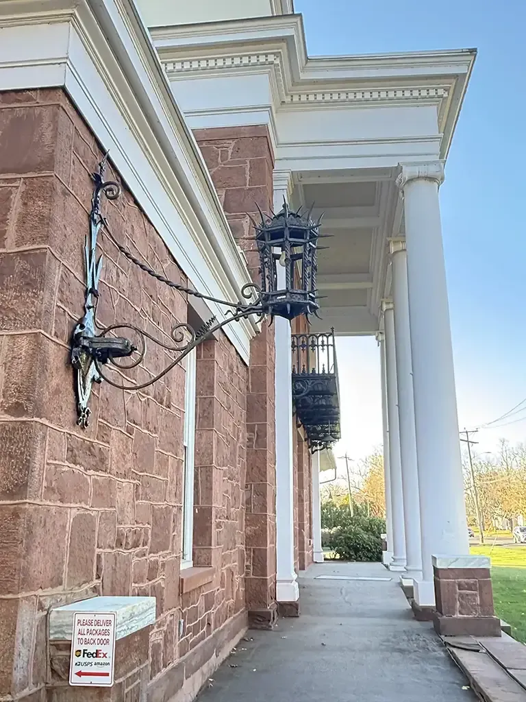 Historic stone building entrance with white columns, mounted cables, and a sidewalk on a sunny day