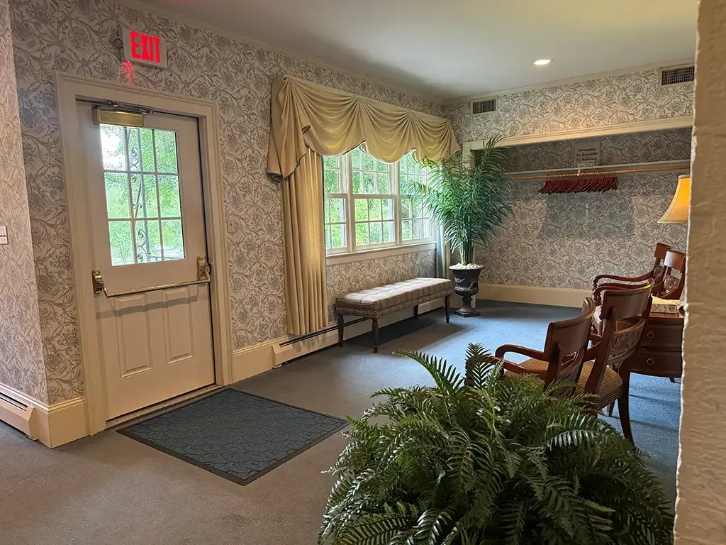 Elegant waiting room with patterned wallpaper, wooden chairs, a bench, and large windows beside a door.