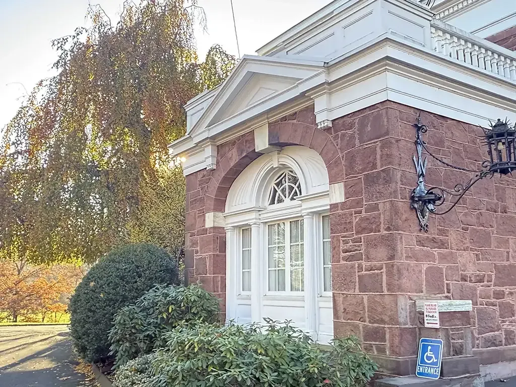 Brick building entrance with white door trim, a hanging lantern, and autumn trees beside a walkway