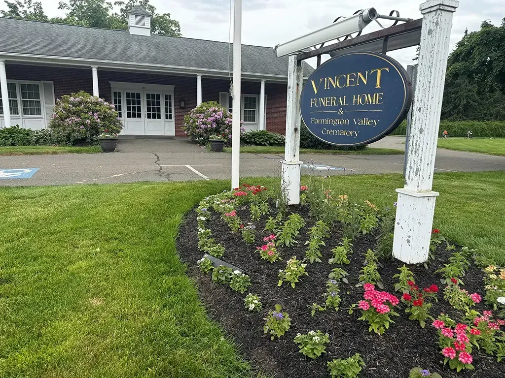 Vincent Funeral Home sign in front of a red brick building, bordered by colorful flower beds and green lawn.