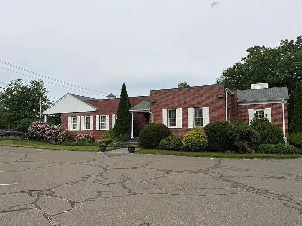 Single-story brick house with white siding and red roof, front shrubs, and cracked driveway under cloudy sky
