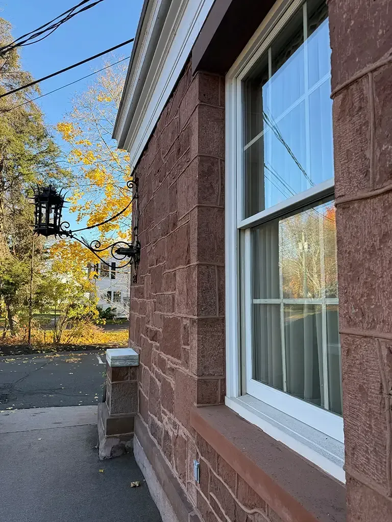 Red brick building wall with a white-framed window beside a sidewalk and autumn trees.