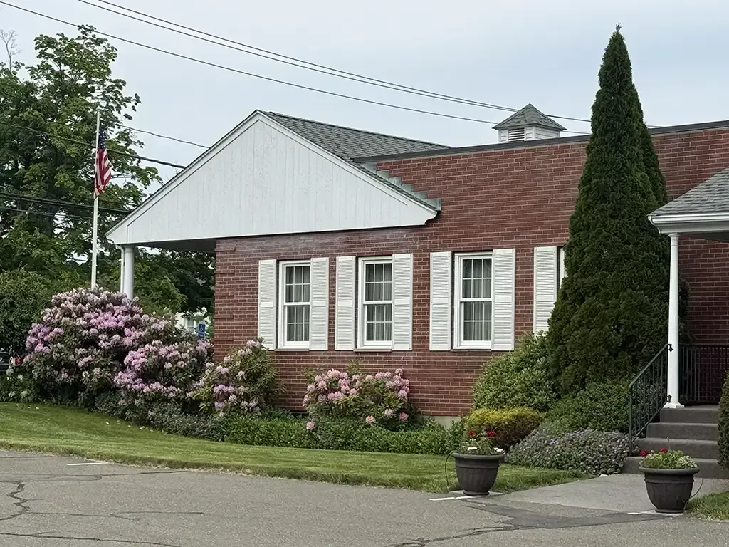 Brick house with white shutters, red roof, and flowering shrubs beside a flagpole