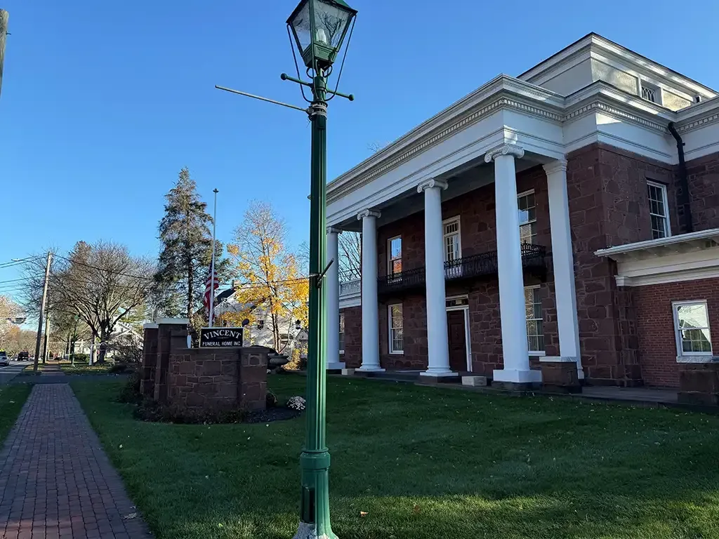 Historic brick building with white columns beside a green lawn and lamppost on a sunny day