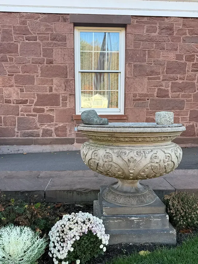 Stone planter in front of a brick wall and window, with white flowers below.