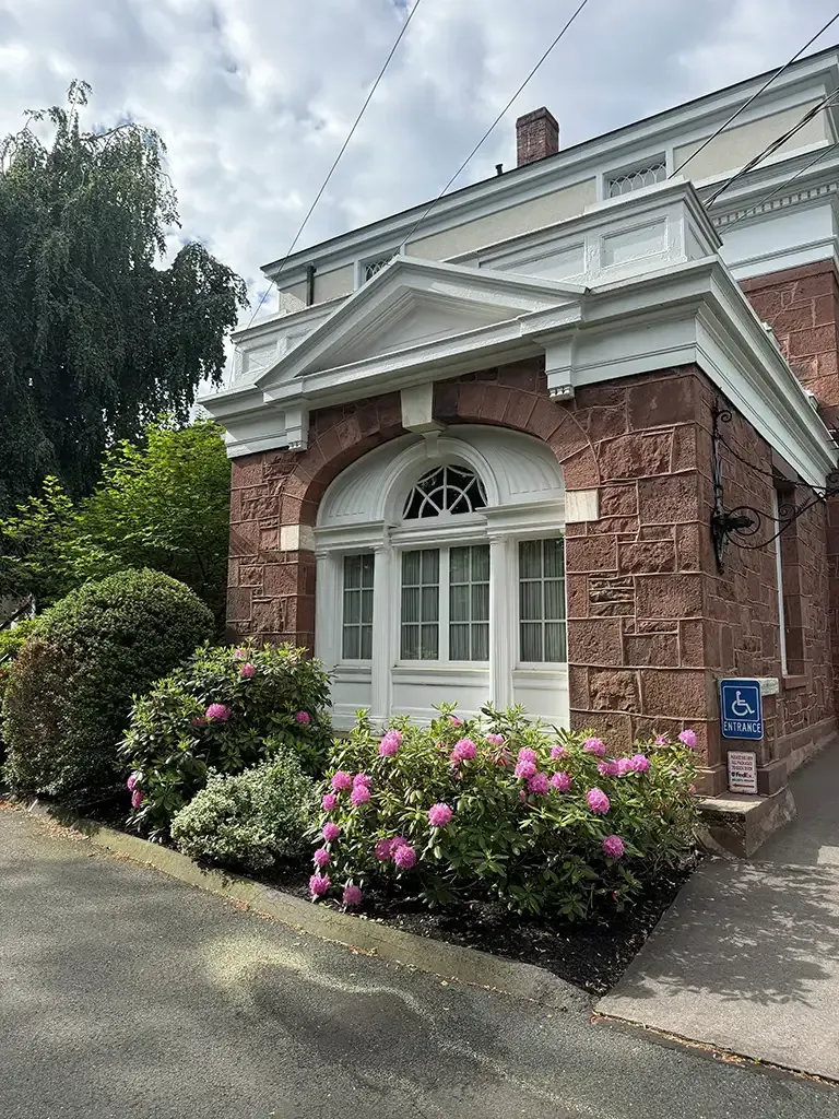 Historic building entrance with white trim, red stone walls, and pink flowers along the walkway
