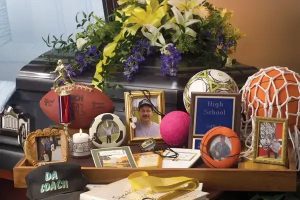 Memorial display with flowers, awards, framed photo, balloons, and a cap reading “Da Coach”