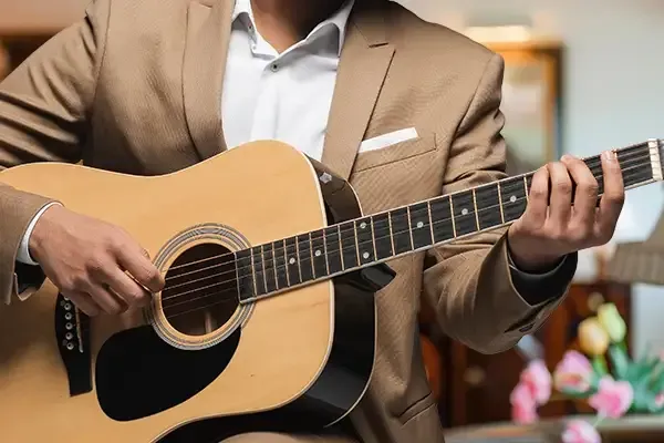 Person in a tan suit playing an acoustic guitar indoors with a blurred floral background