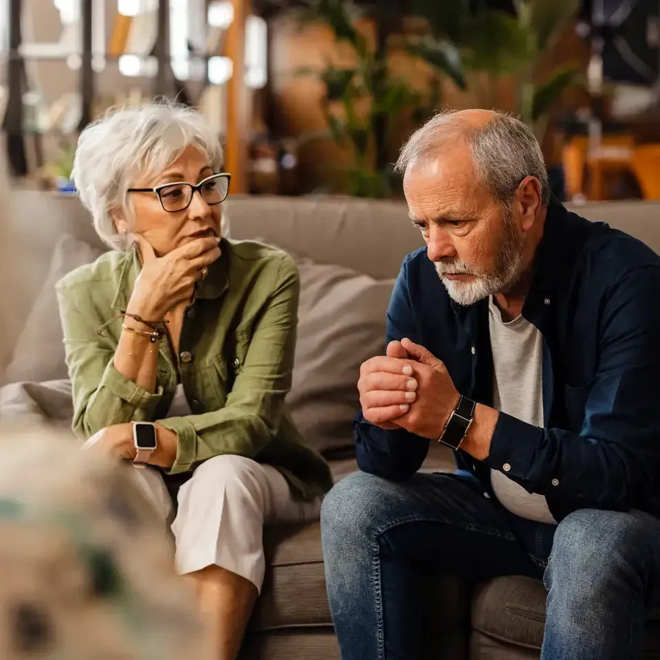 Two people sit on a couch, talking thoughtfully in a warm living room.