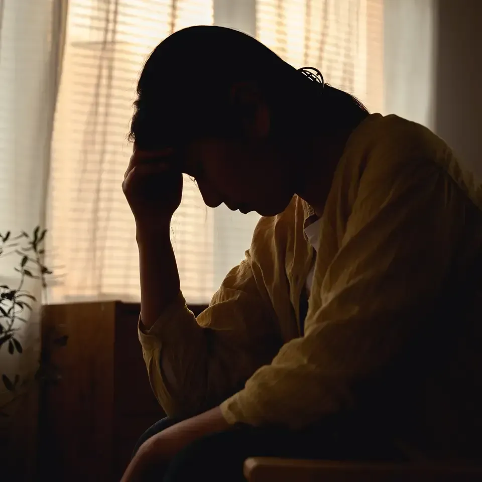 Silhouetted person sitting by a window, hand on forehead, in a dimly lit room