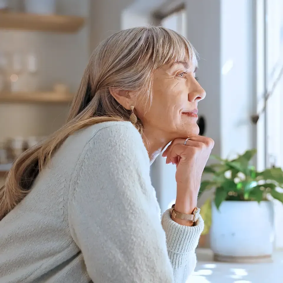 Thoughtful woman by a window, resting her chin on her hand beside a potted plant