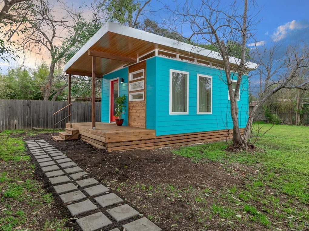 Teal tiny house with red door, wooden porch, and walkway on a grassy lawn.