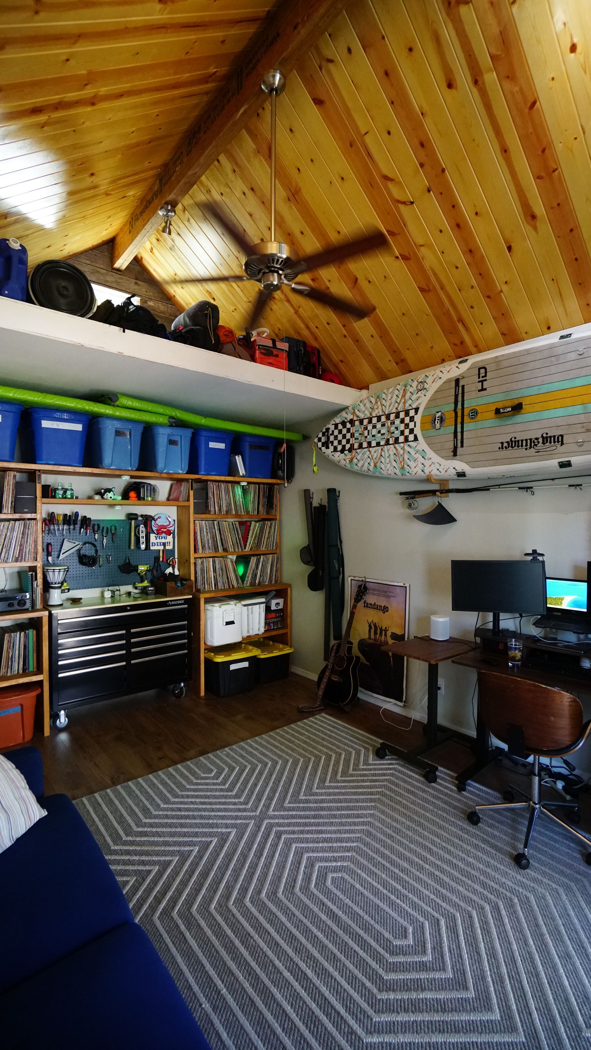 Interior view of a loft-style room with a wooden ceiling and a desk, surfboard, and storage.