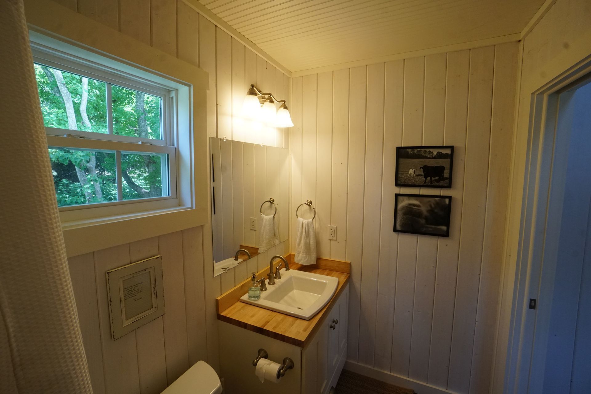 Small white bathroom with a window, sink, mirror, and two framed pictures on the wall.