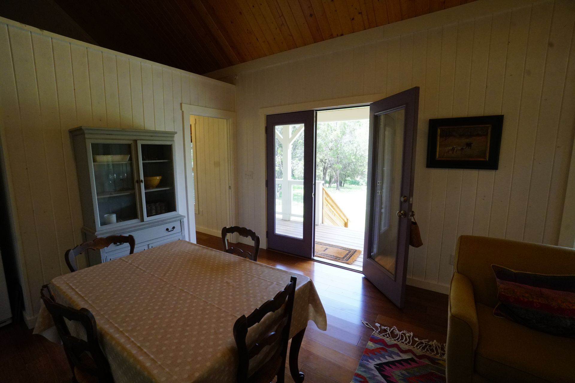 Dining room with table, chairs, hutch, and open purple-framed doors to a porch; white walls, wooden ceiling, rug.