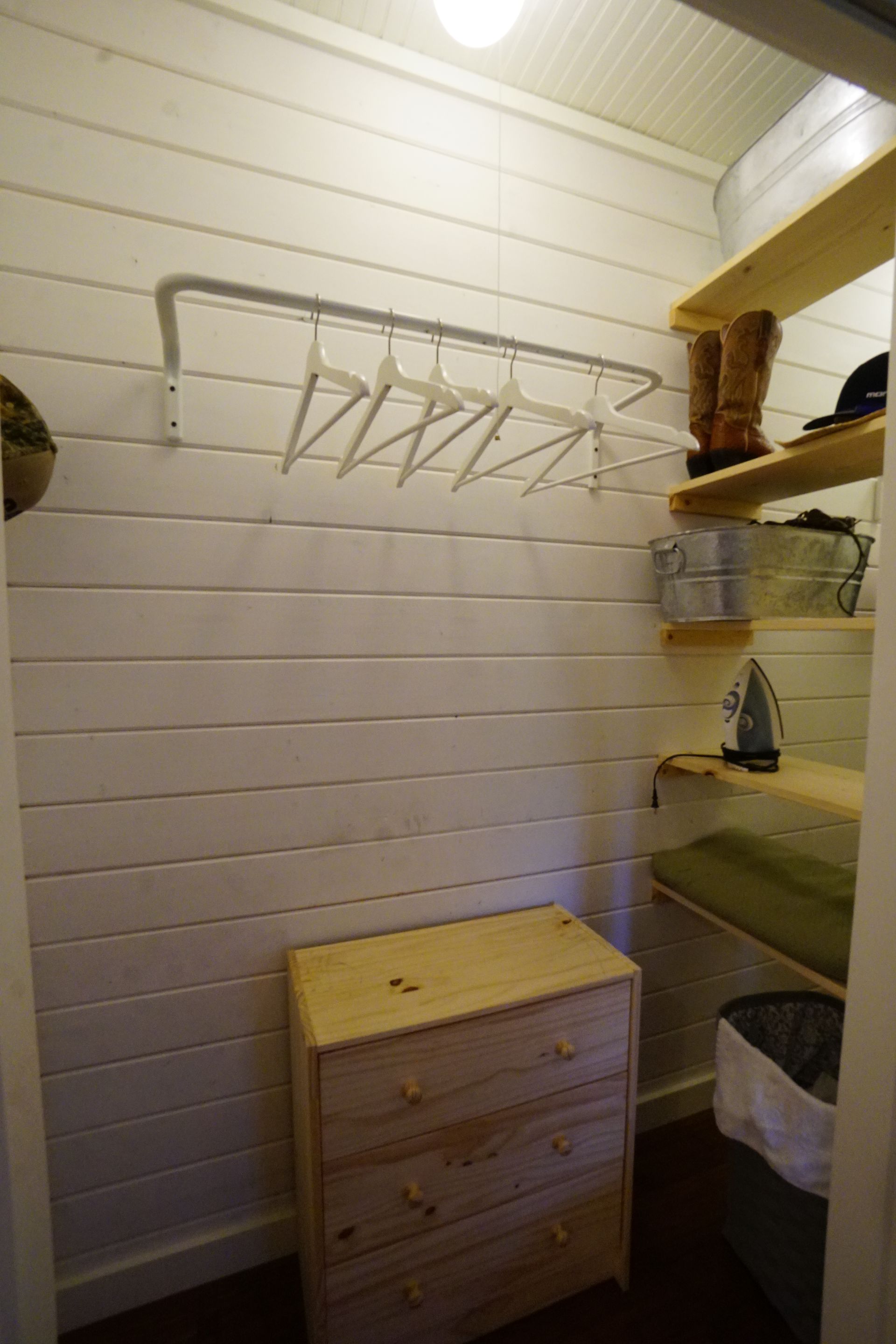 Small closet with white horizontal wood paneling, clothes rack, wooden shelves, and a chest of drawers.