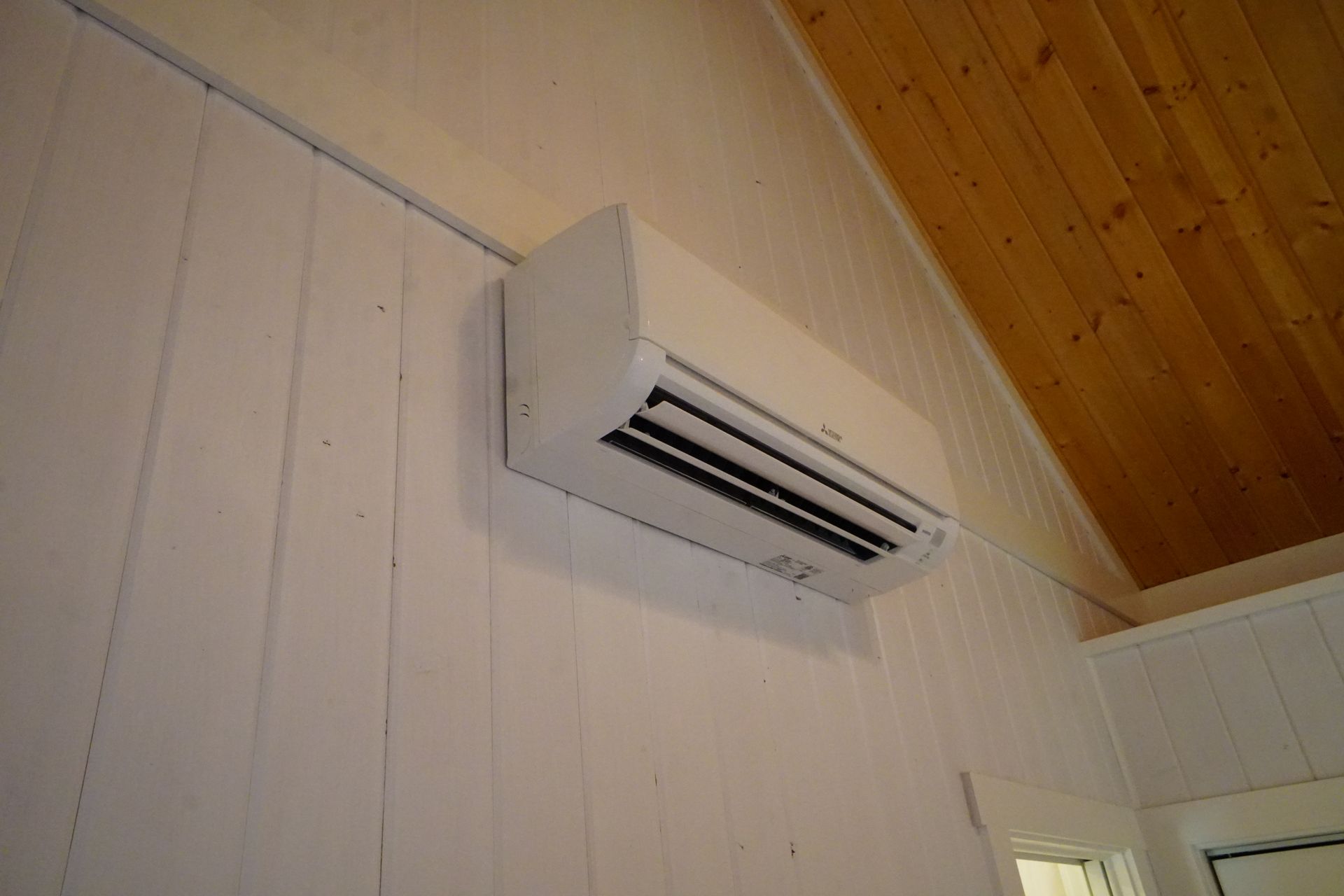 White wall-mounted air conditioner in a room with a white wall and wooden ceiling.