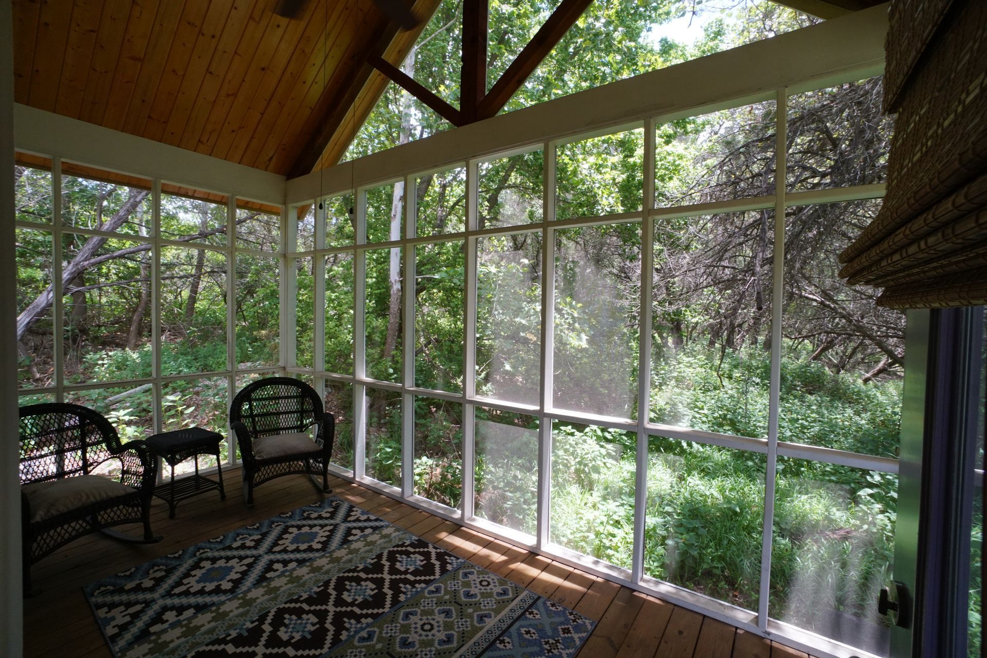 Screened-in porch with wicker furniture and rug, overlooking a lush green forest, natural light.