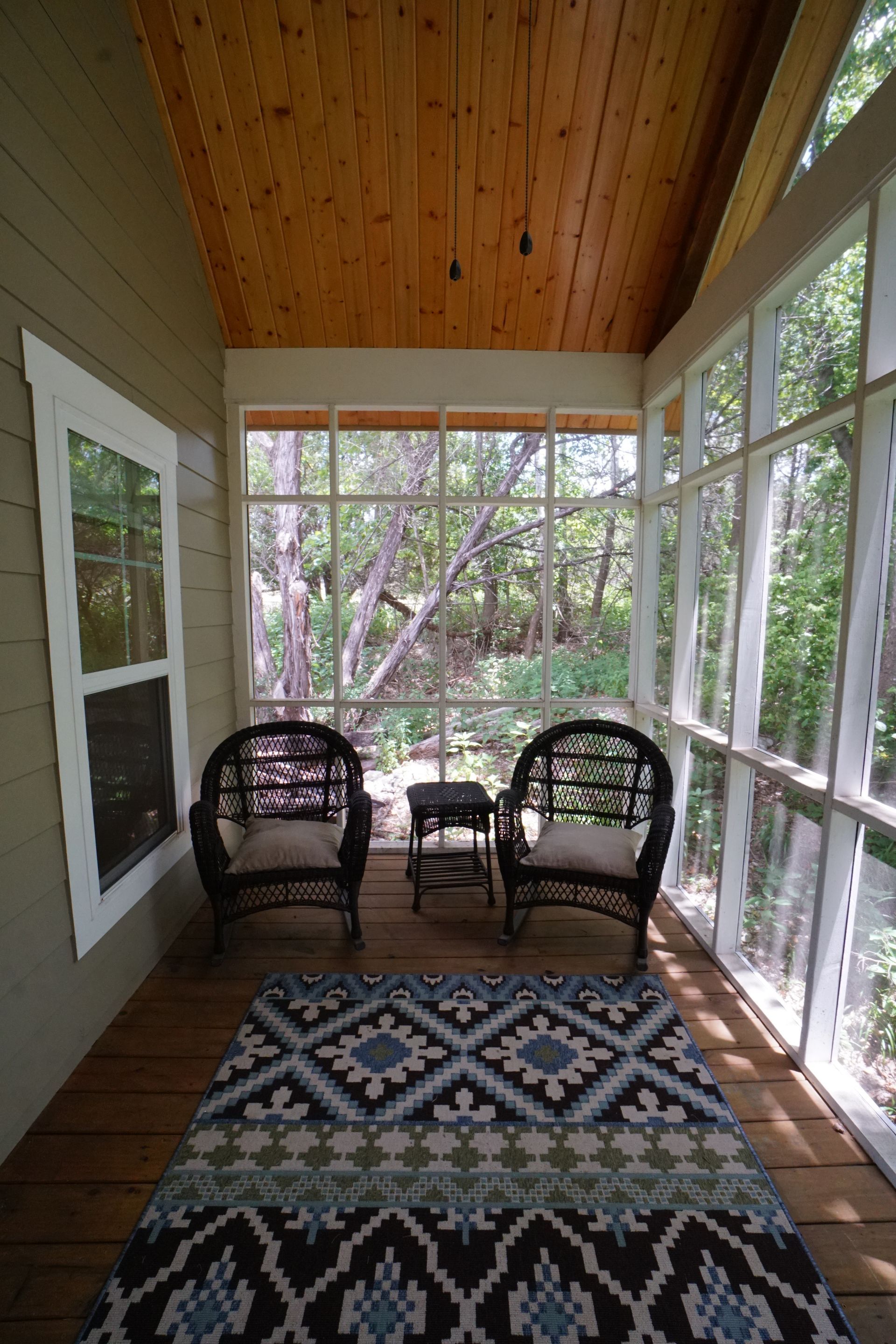 Screened porch with two wicker chairs, small table, patterned rug, and view of trees.