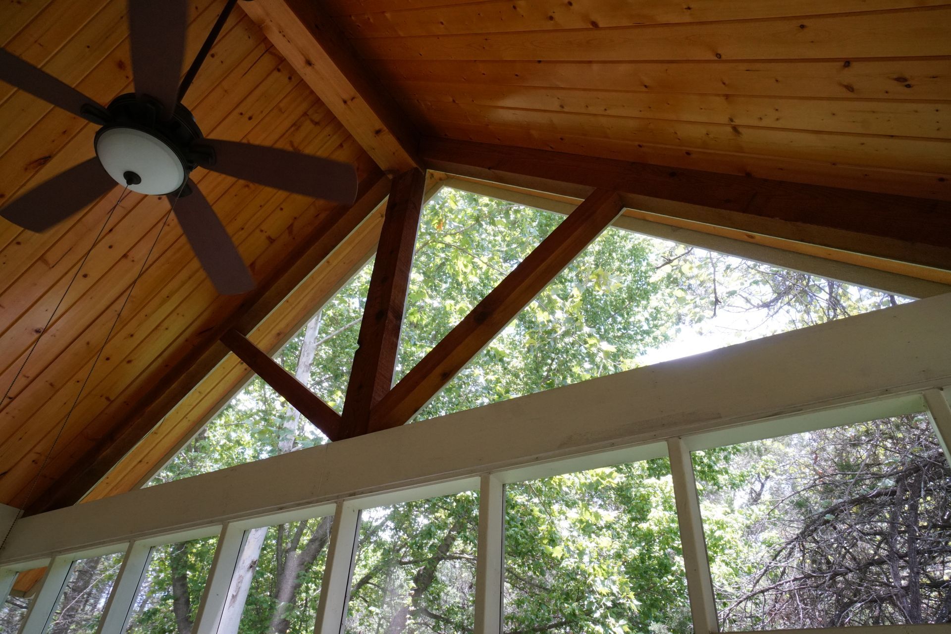 Wooden screened porch ceiling with triangle window, beams, and ceiling fan, overlooking trees.