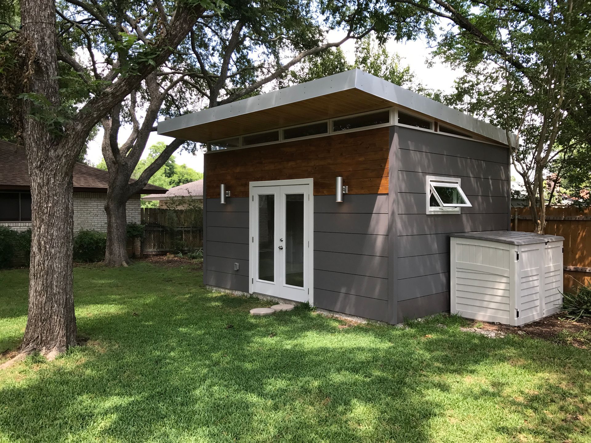 Modern gray and wood shed with double doors, small window, and air conditioner, in a green yard.