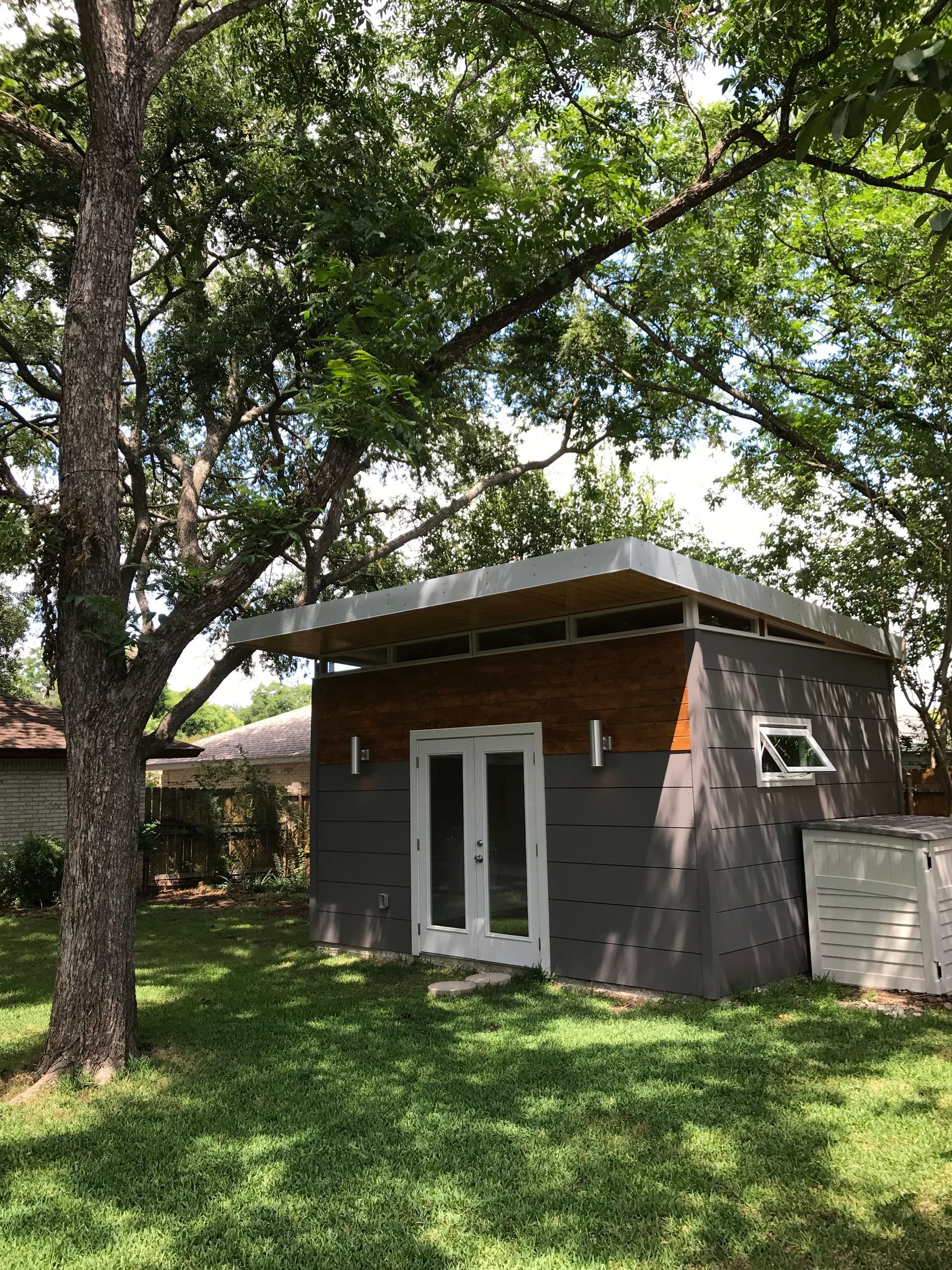 Modern backyard studio with gray siding, wood accents, and double doors, beneath tree branches.