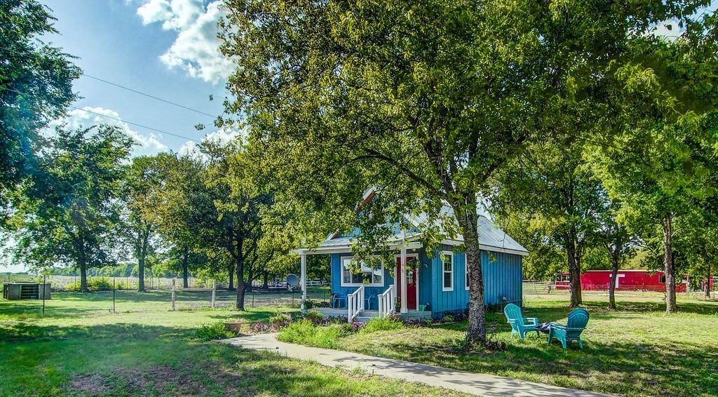 Blue cottage with red door under tree, chairs on lawn, sunny day.