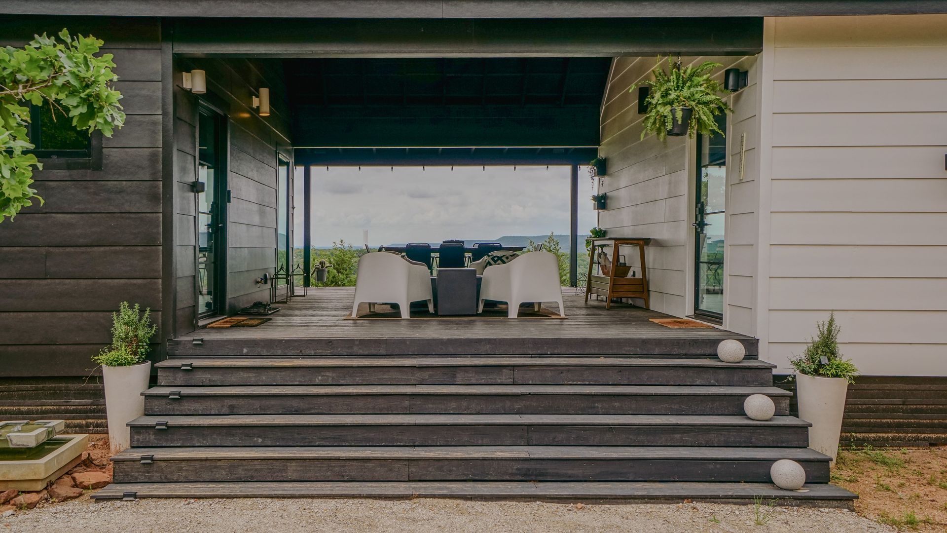 Stone steps lead to a porch with white chairs and a table overlooking a landscape.