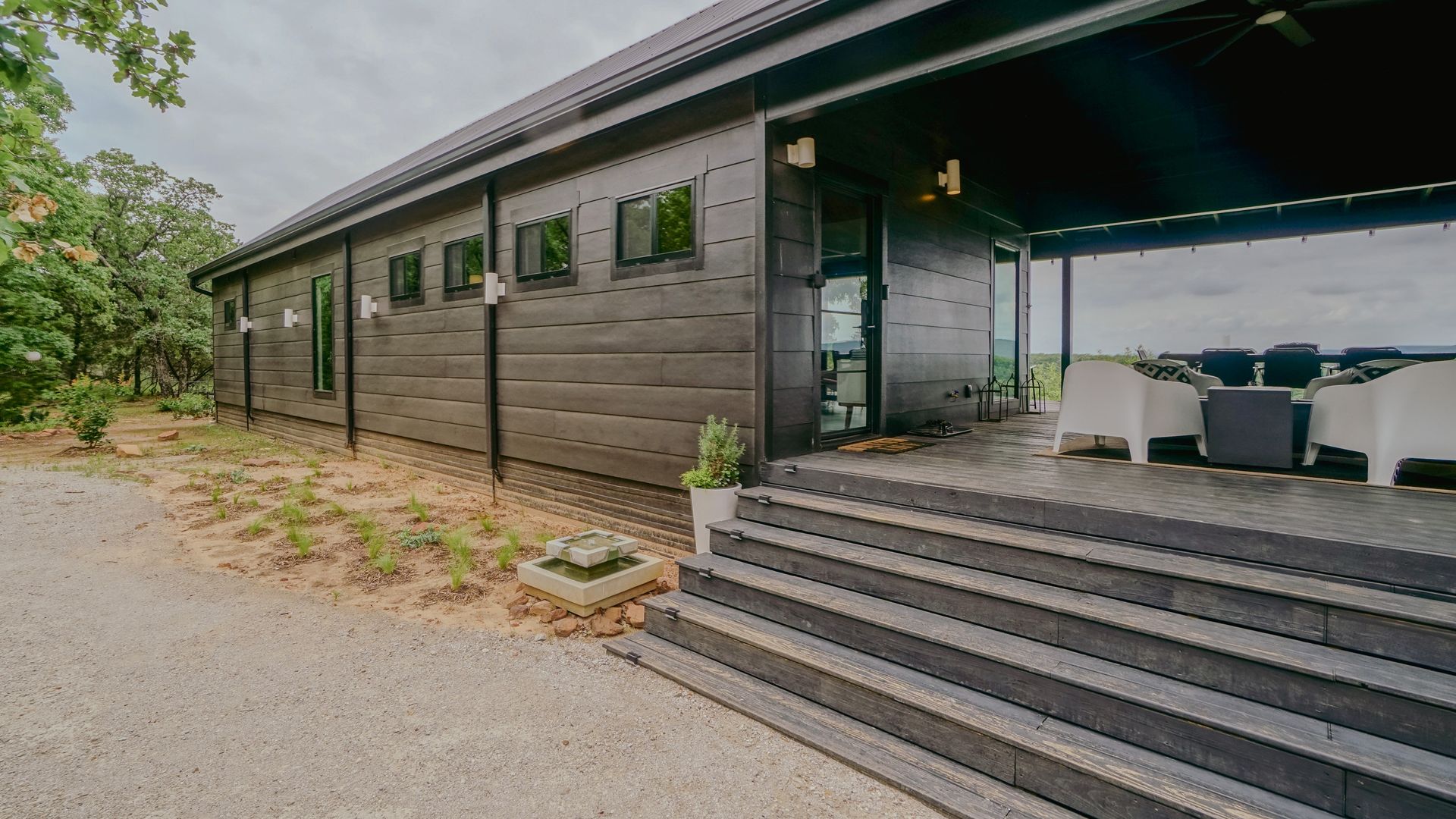 Dark-colored building with steps leading to a covered patio with white furniture, set on a gravel driveway.