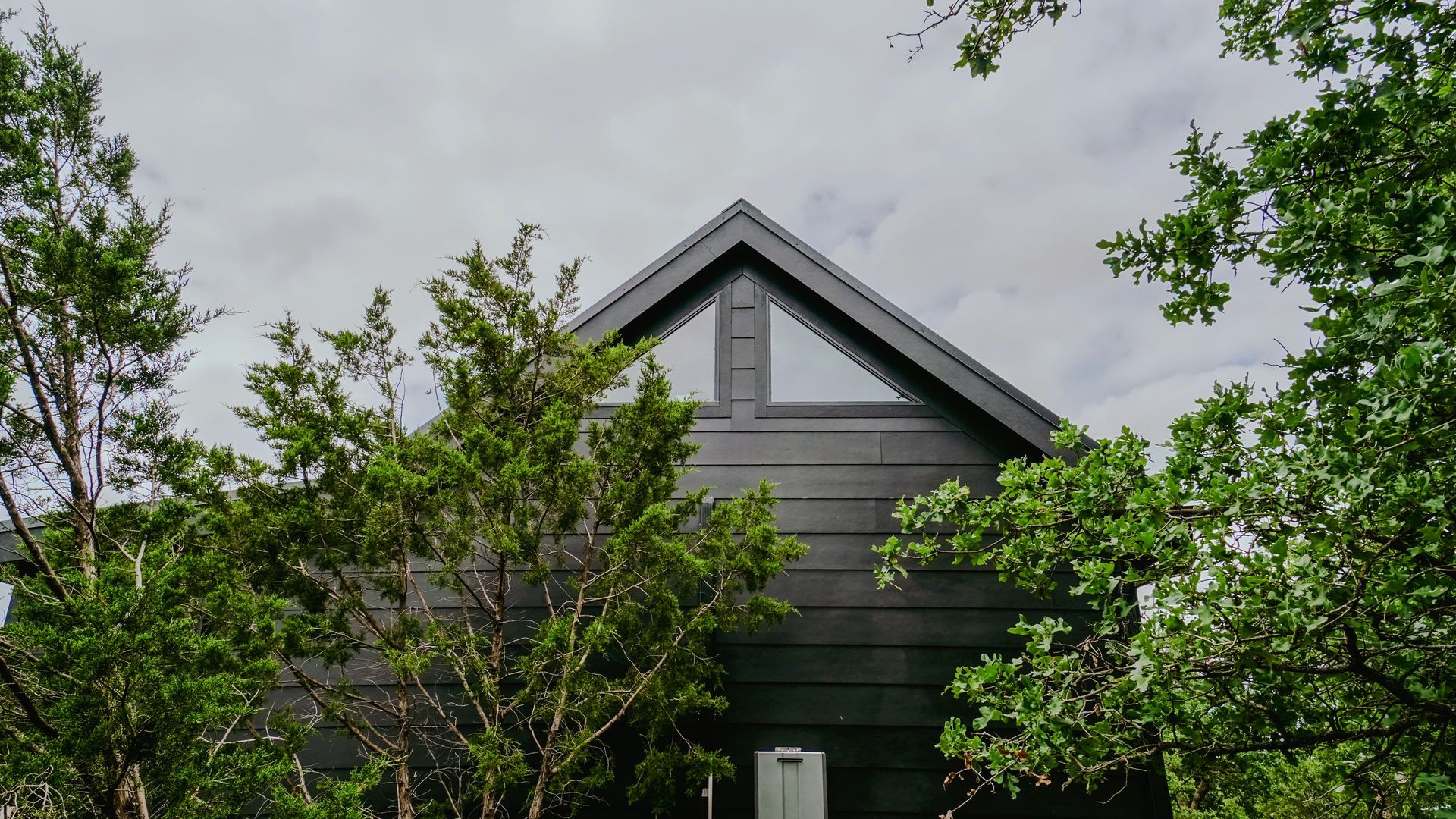 Dark gray cabin exterior with triangular window, framed by green trees against a cloudy sky.