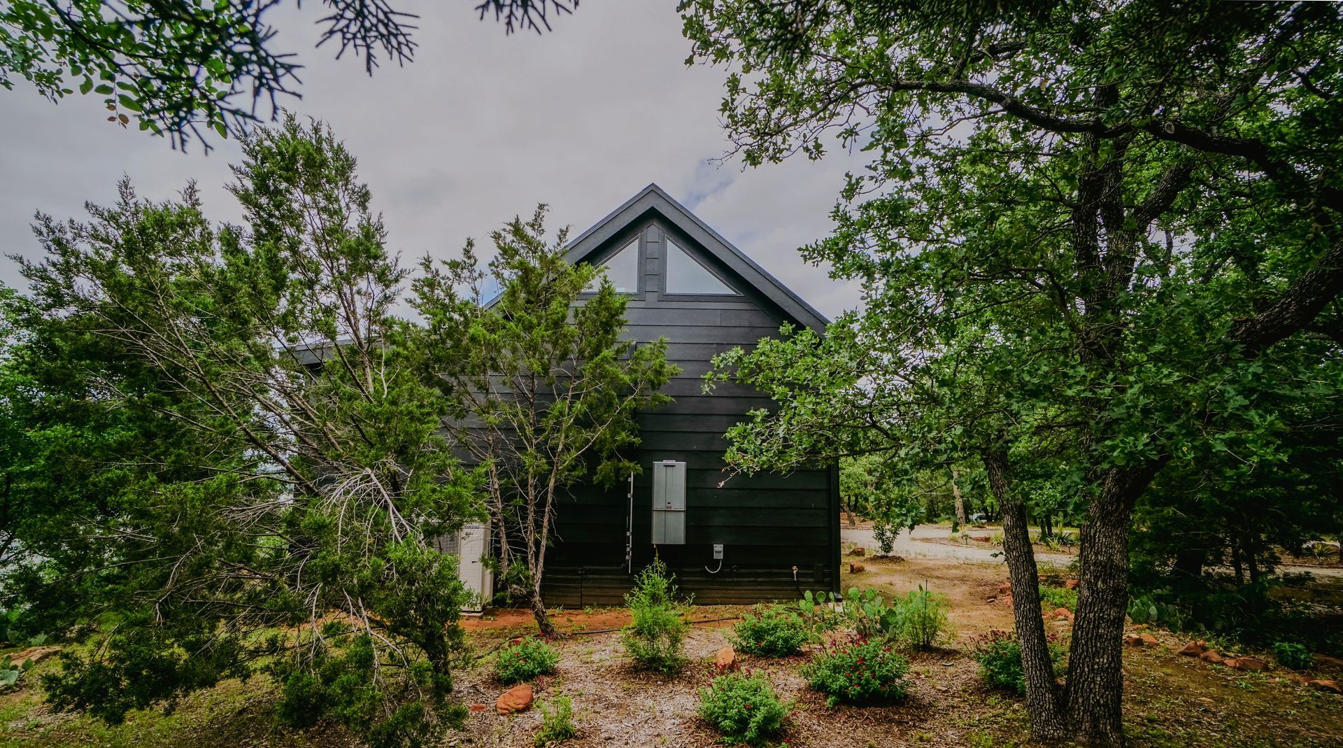 Black cabin with triangular roof, nestled among green trees.