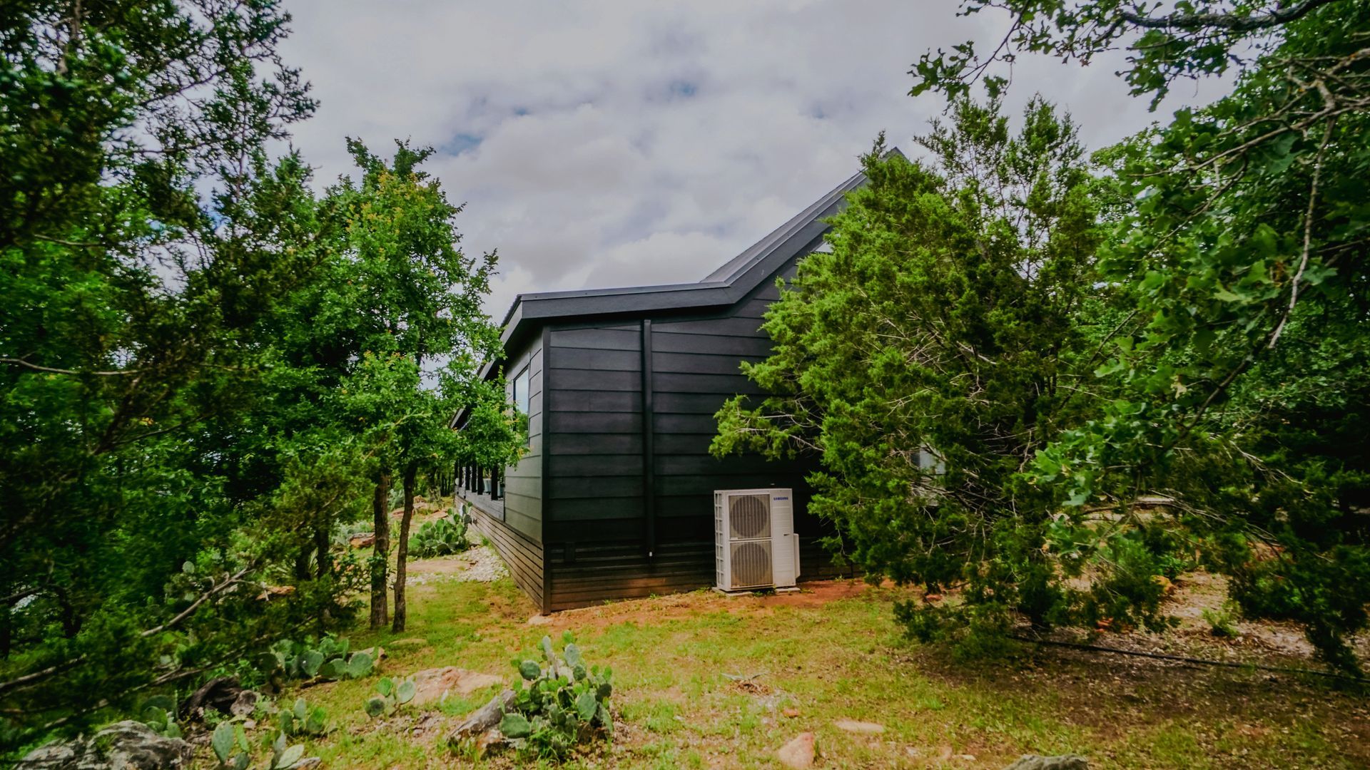 Black-sided cabin nestled in greenery with an air conditioner unit on the side. Cloudy sky overhead.