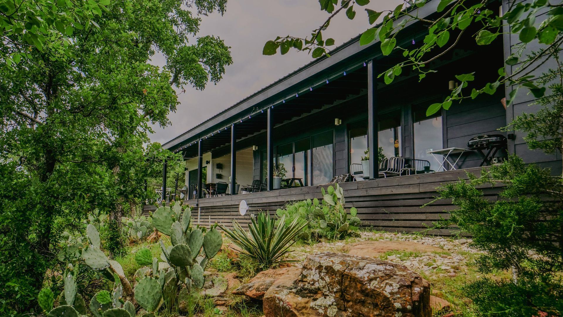 Black modern house with a long porch, surrounded by greenery and cacti.