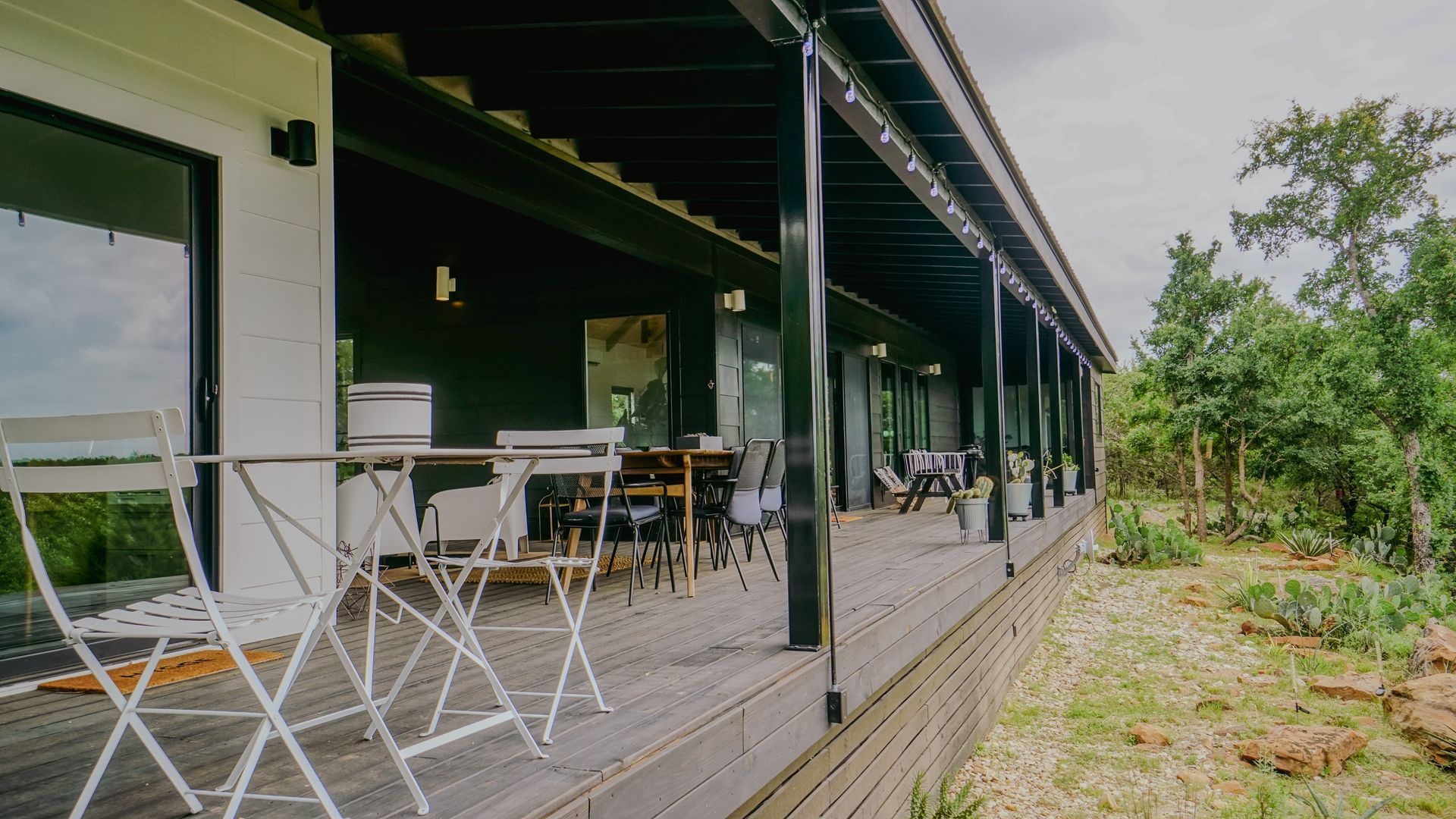 Long, covered wooden deck with tables and chairs outside a building, surrounded by greenery.