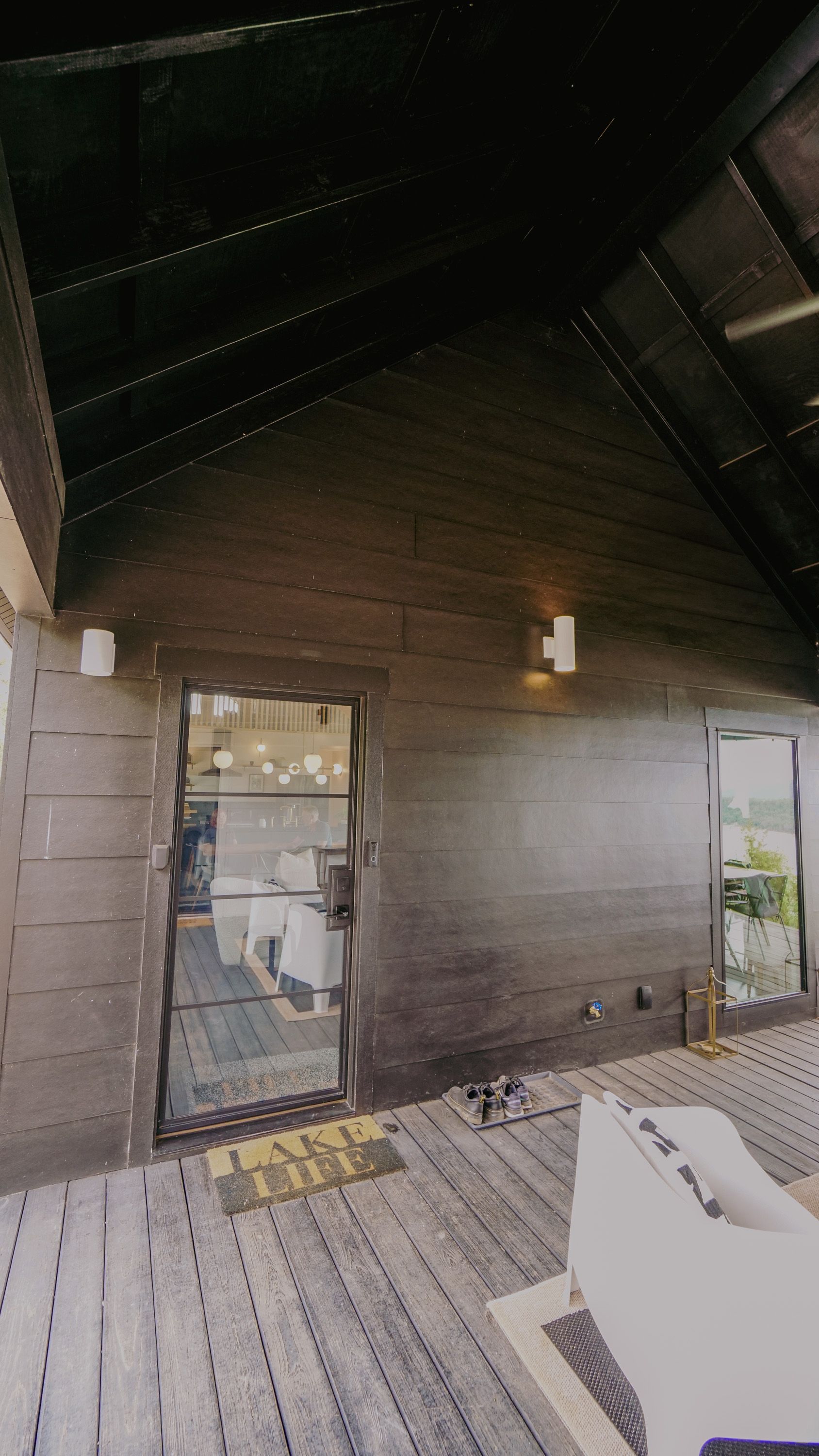 Exterior view of a modern cabin. Dark wooden walls, glass doors, and a gray deck with outdoor furniture.
