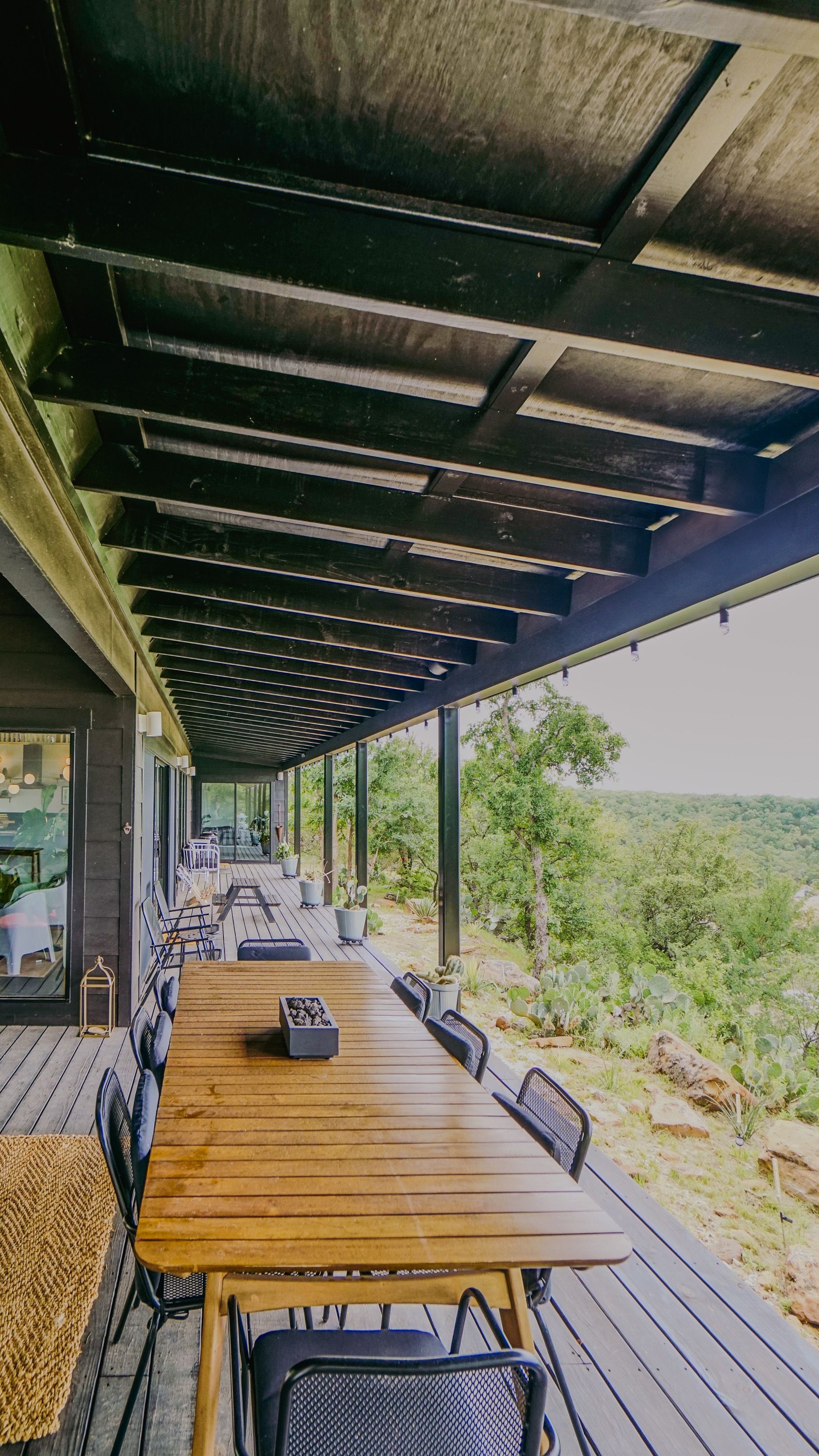 Long outdoor dining table on a deck with forest views, beneath a dark wood ceiling.