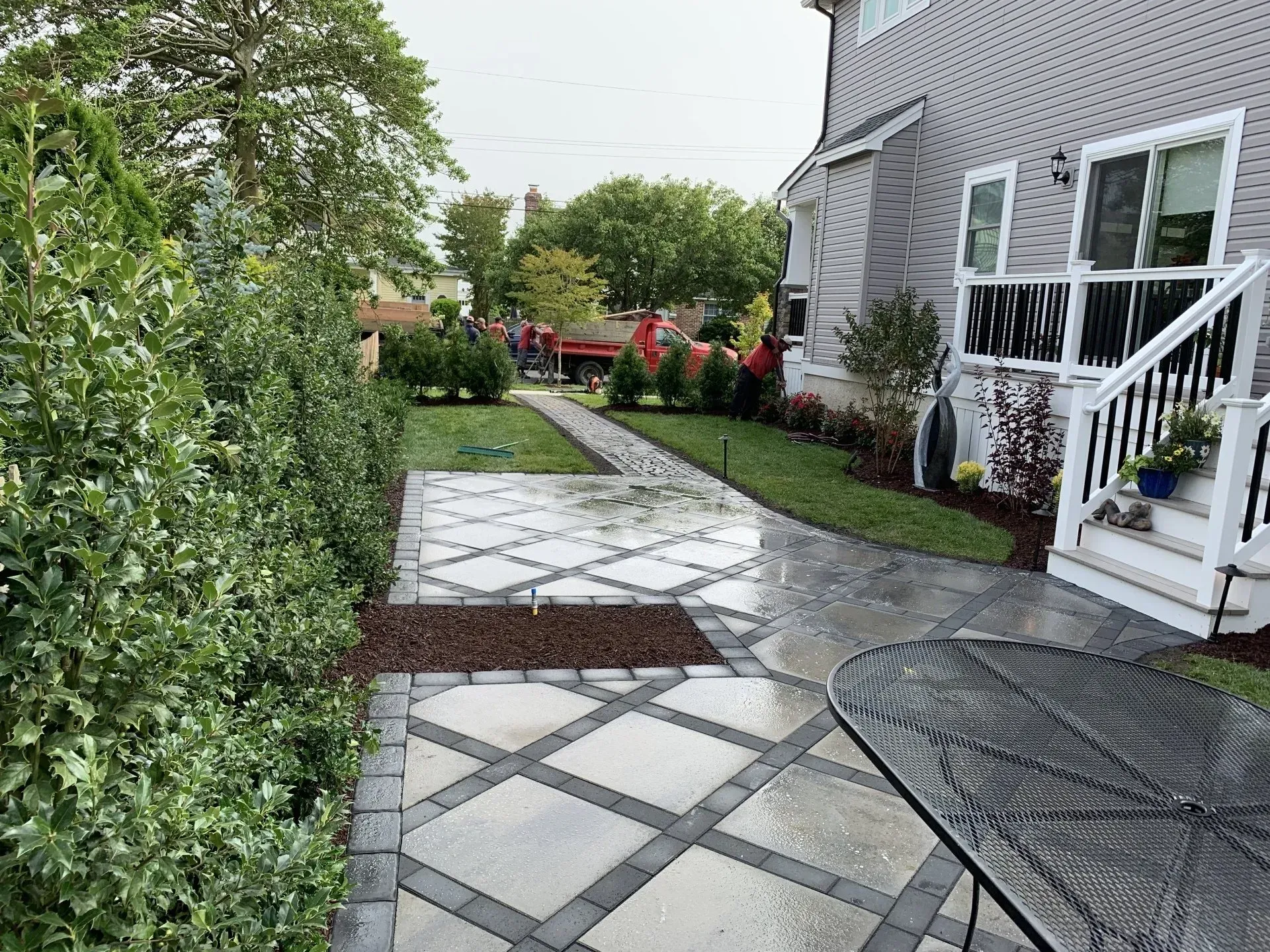 Curved stone patio path beside a house, bordered by hedges and potted flowers, leading to a backyard garden.