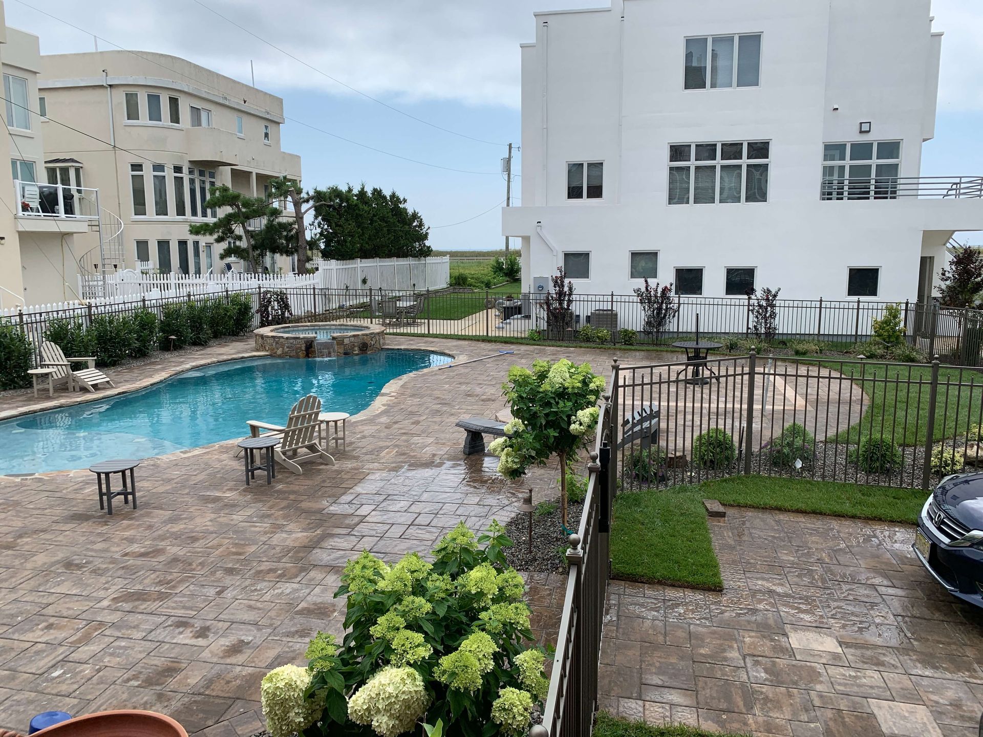 Swimming pool courtyard between white apartment buildings, with patio chairs, greenery, and a parked car.
