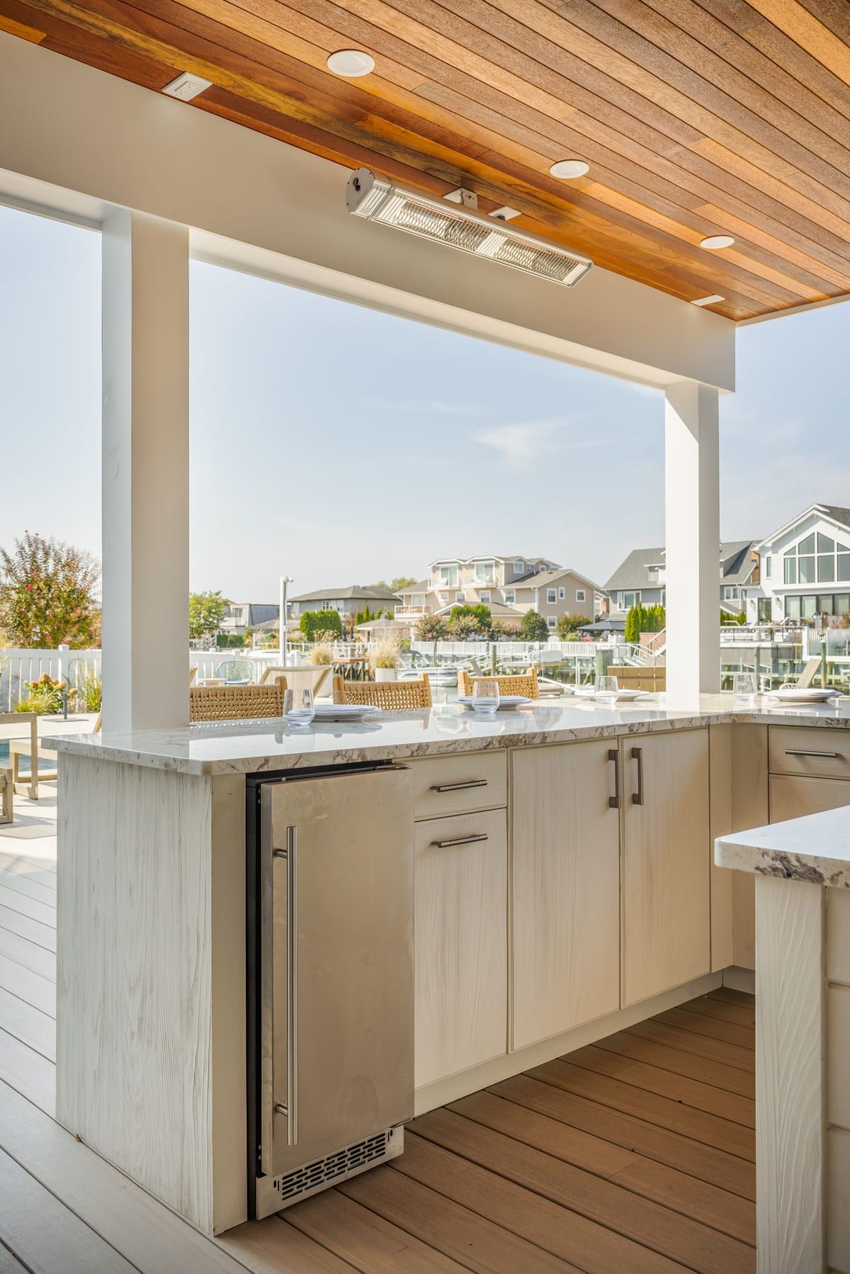 Outdoor kitchen with sink, cabinets, and countertop under a wood ceiling, overlooking a waterfront neighborhood