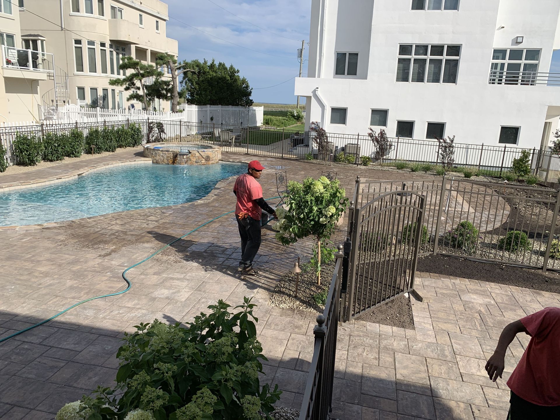Person in red shirt watering plants beside a small pool in a courtyard with white buildings.