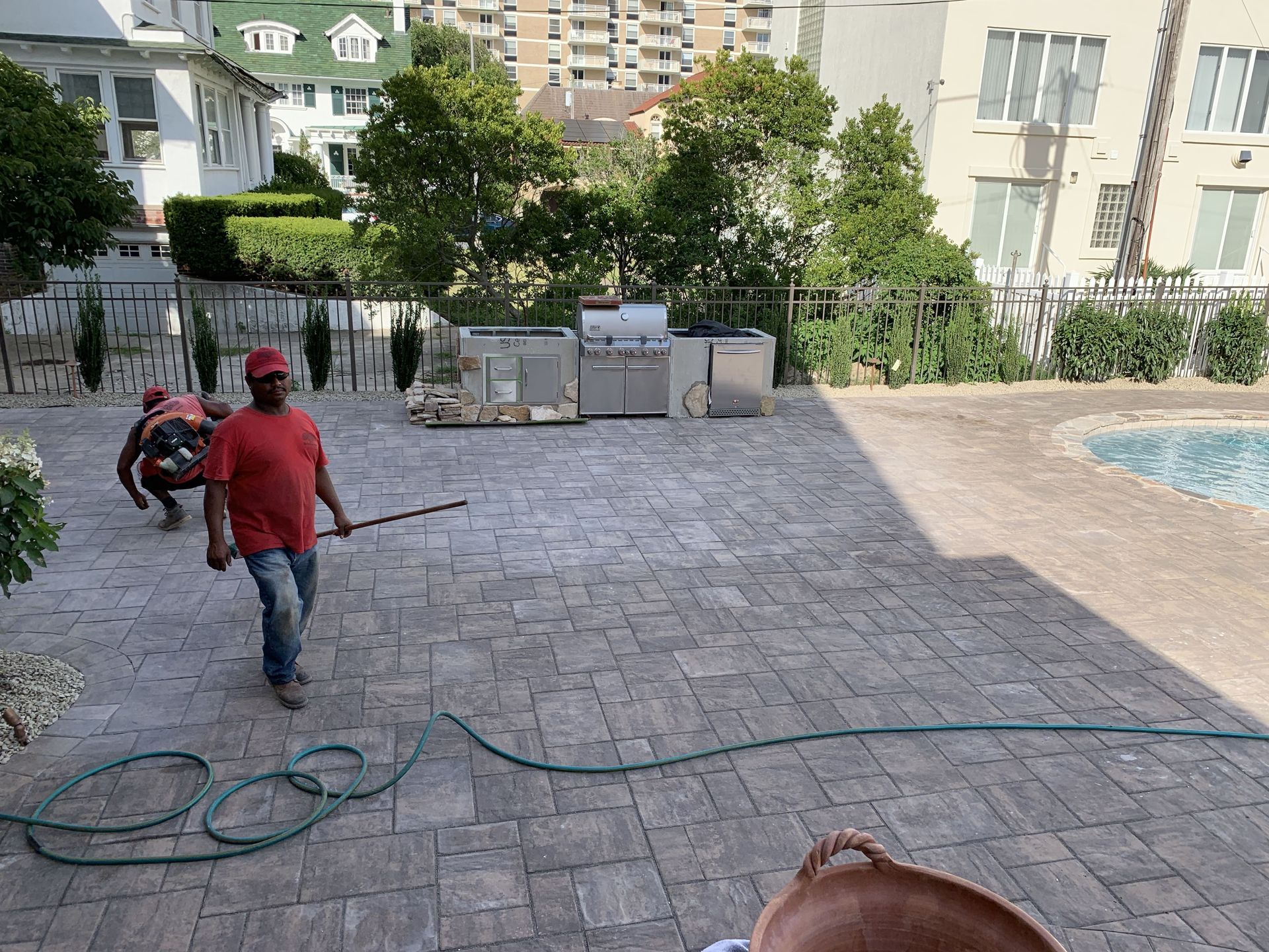 Two workers on a stone patio with a coiled hose, pool at right, and apartment buildings in the background