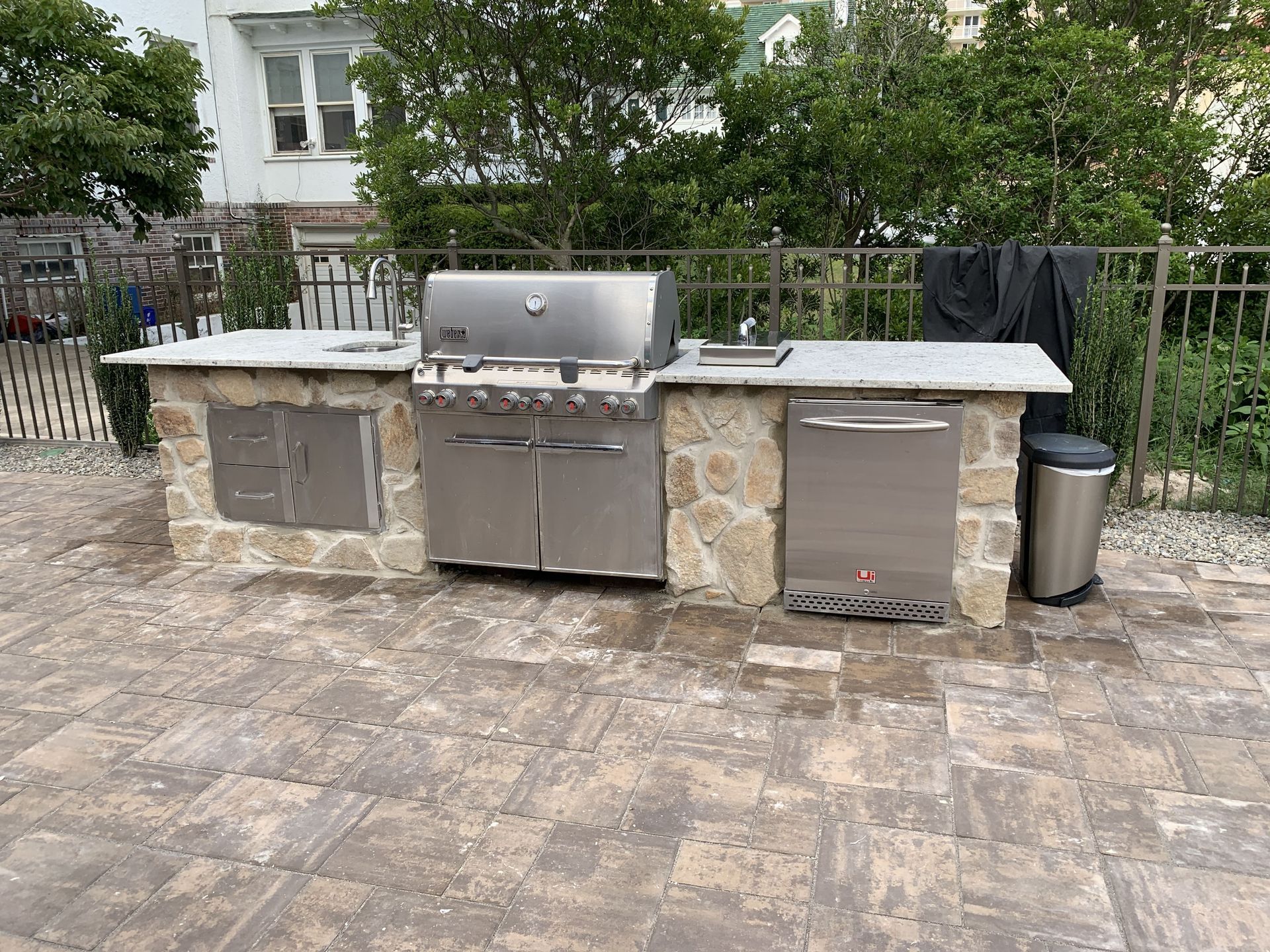 Outdoor stone patio with a built-in stainless steel grill and side counters, backed by a metal fence and shrubs