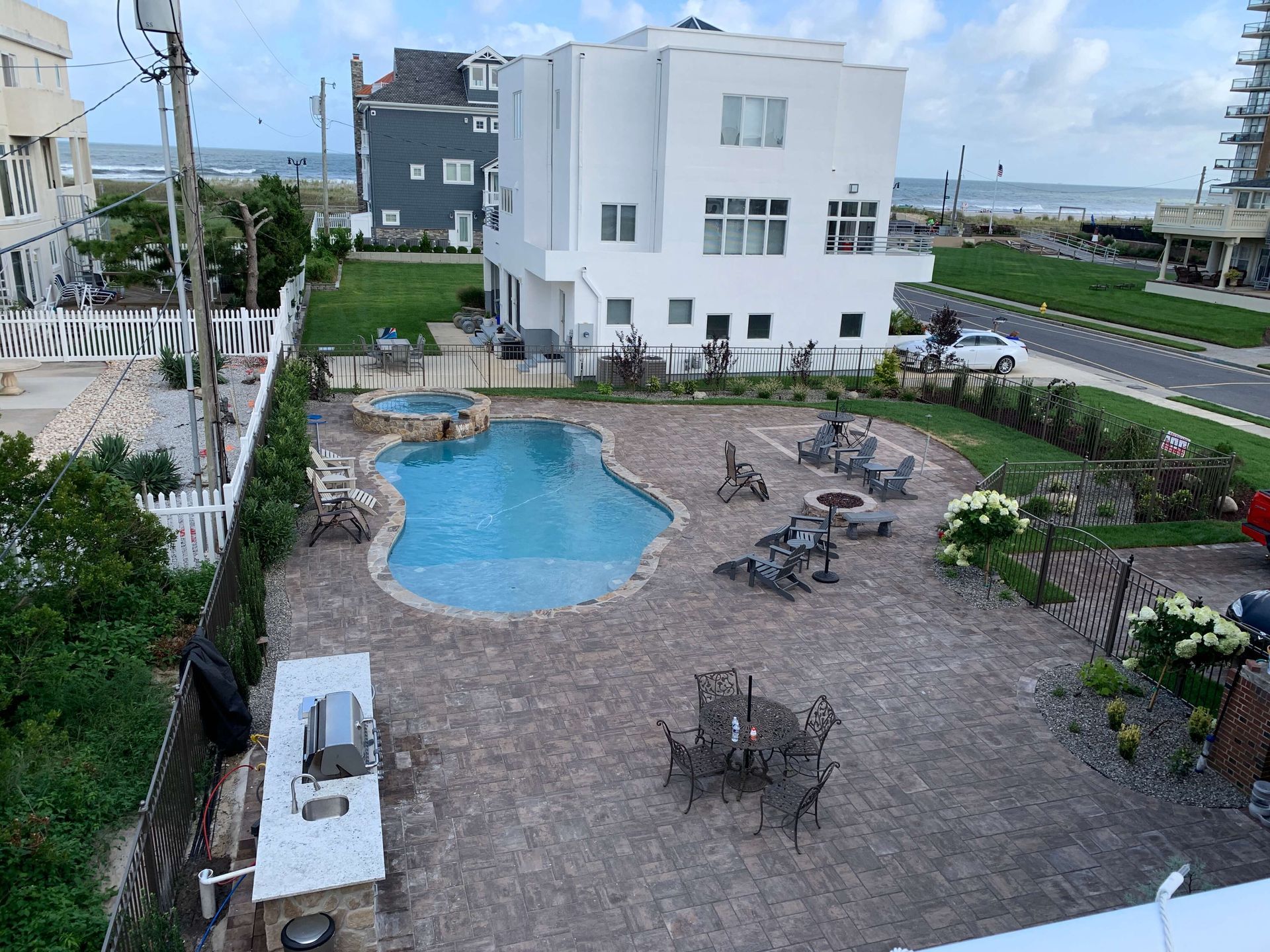 White modern house beside a small blue pool and patio with outdoor tables, surrounded by greenery