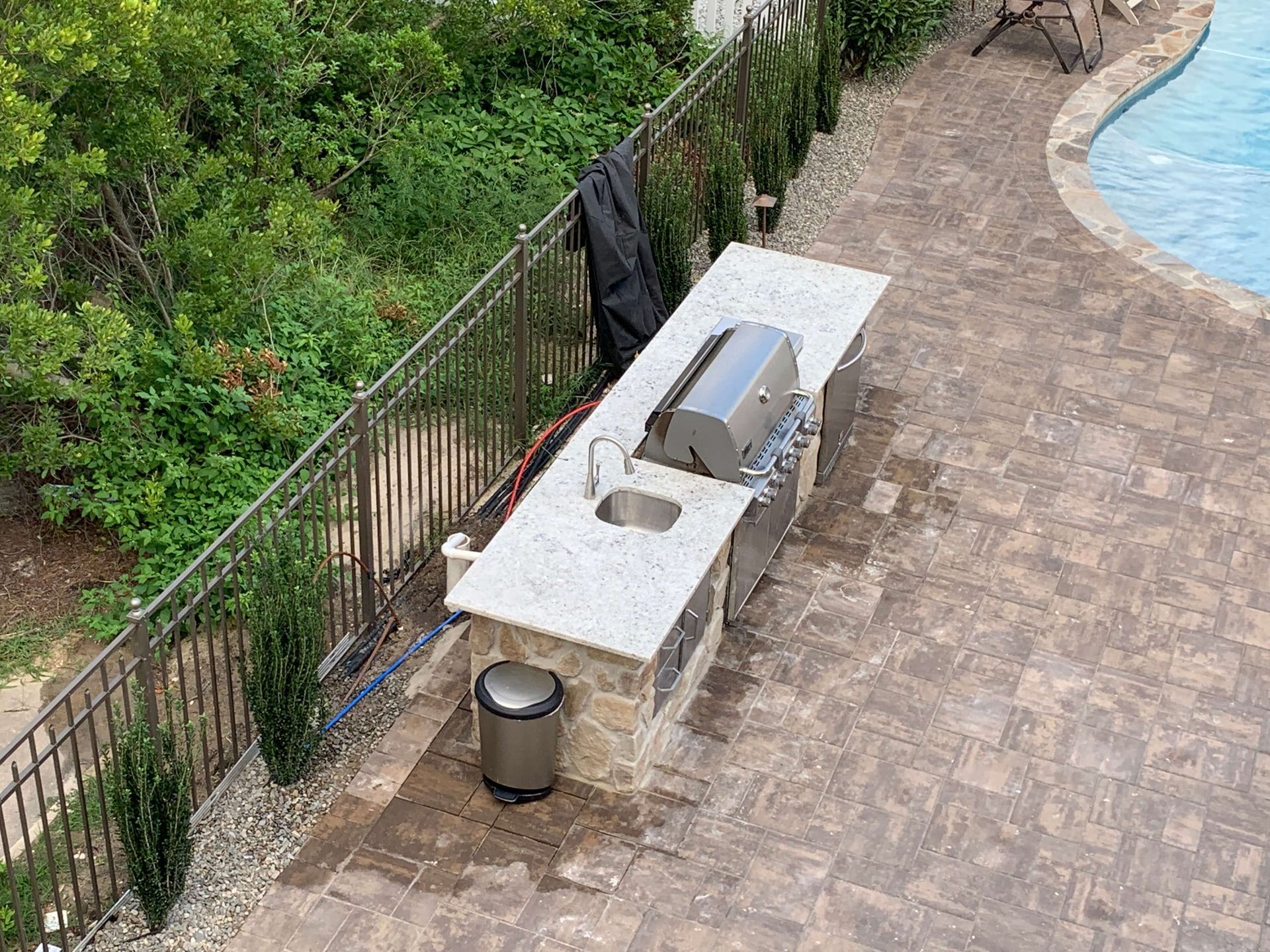 Outdoor poolside walkway with a white bench, trash can, and black towel hanging on a fence рядом