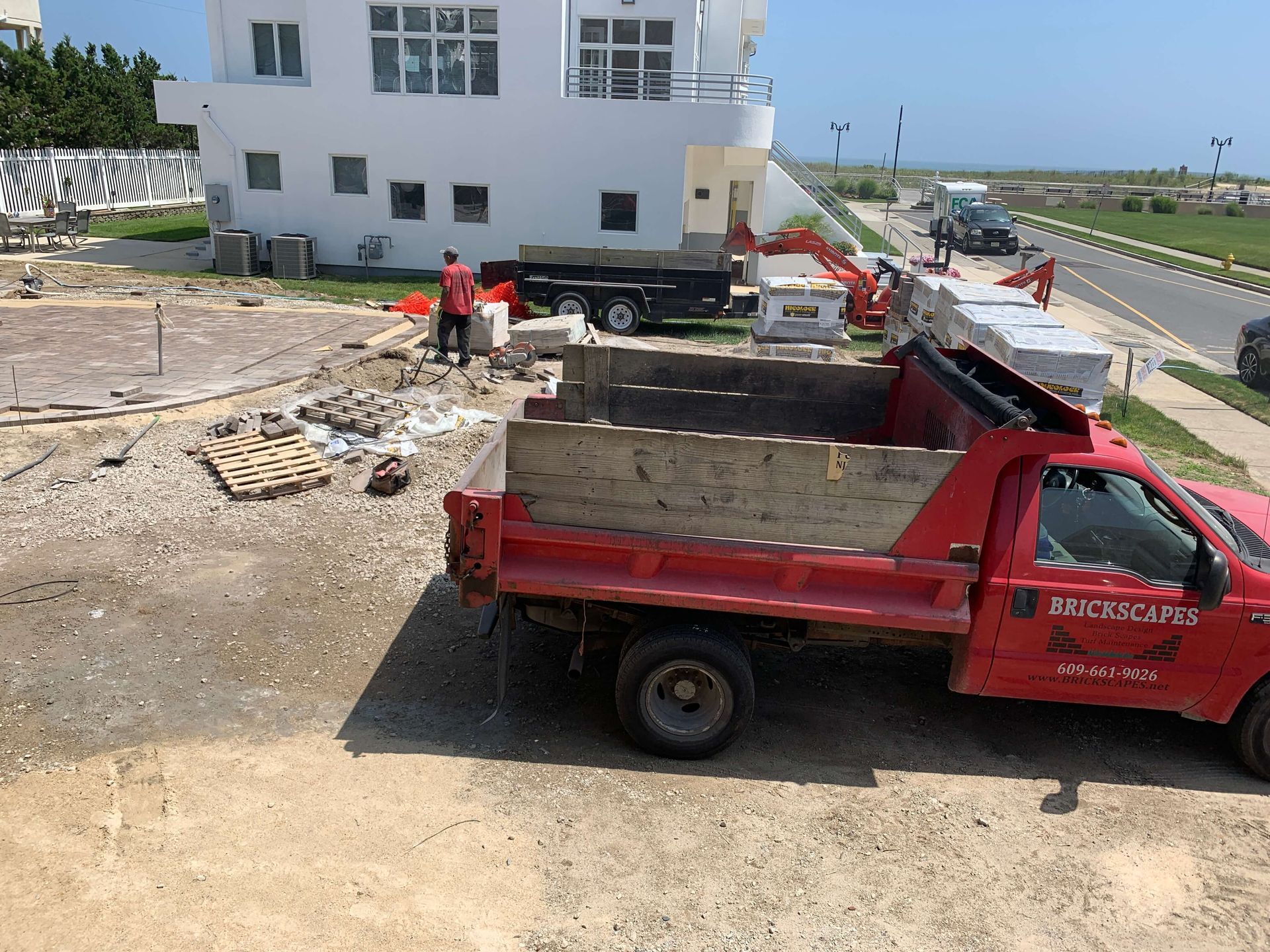Red dump truck and excavator at a construction site beside a white building and road