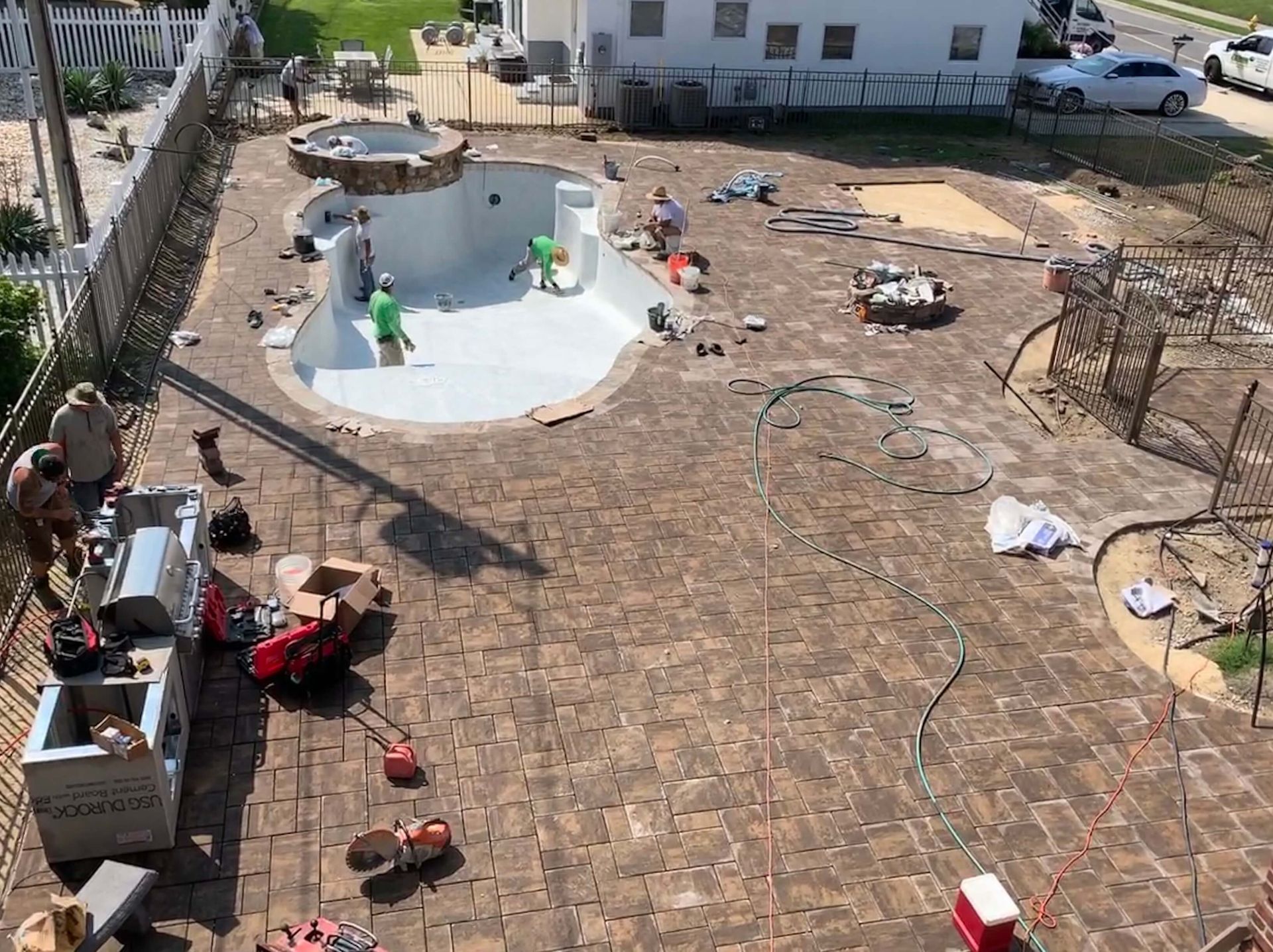 Backyard under construction with a white pool, workers, tools, and a stone patio laid out around it